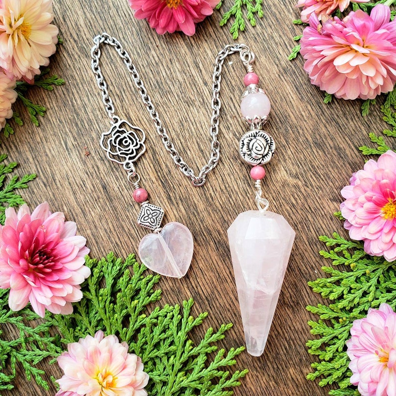 A pink rose quartz crystal pendulum with a silver chain and rose-shaped charms. The beaded chain has pink Rhodonite beads and a rose quartz heart bead at the end.