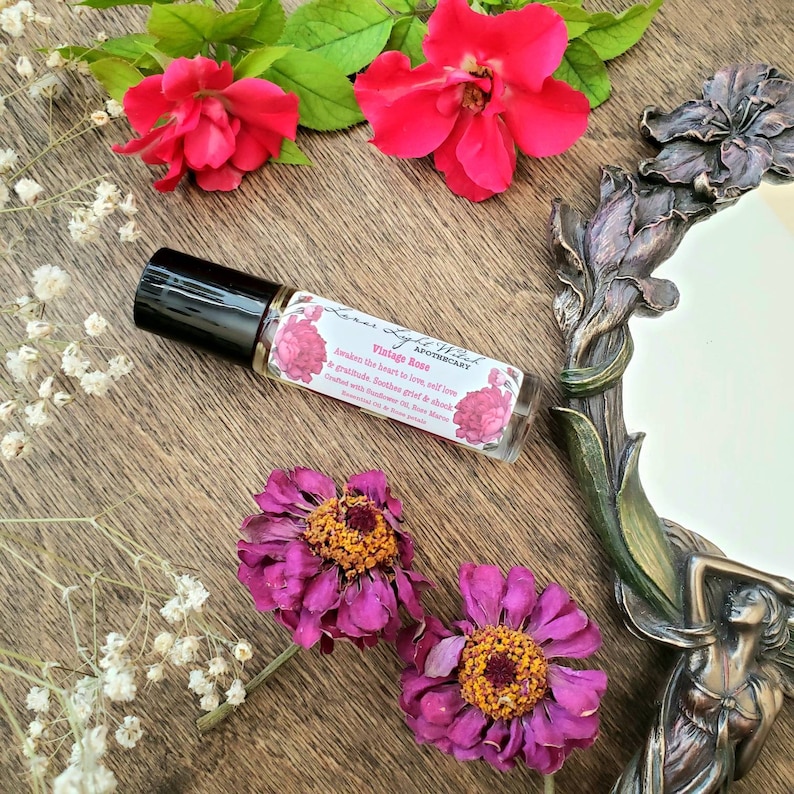 Op de afbeelding: Een klein glazen flesje met een zwarte rolbal. Het etiket is wit met roze bloemen en zwarte tekst die "Light Wilk Apothecary Vintage Rose Awaken the heart to love, self love & gratitude. Soothes grief & shock. Crafted with  Rose Absolute & other  Essential Oils. Rose  Aromas." luidt.