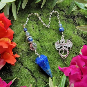 Puede incluir: Un péndulo de cristal de lapislázuli azul con una cadena de plata y un dije de dragón de plata. El péndulo está rodeado de musgo verde y flores rosas.