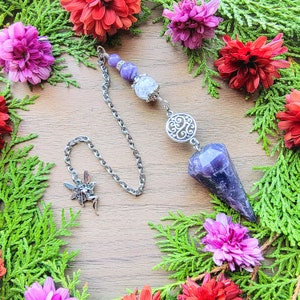 May include: A purple amethyst crystal pendulum with a silver chain and a silver charm. The pendulum is surrounded by purple and pink flowers.