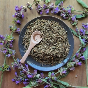 May include: A wooden spoon with a crescent moon and stars design sits on a blue plate filled with dried herbs. The plate is surrounded by purple sage flowers.
