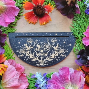 May include: A black wooden half-moon shaped wall decor with a white floral design and a skull in the center. The design is surrounded by a scattering of white stars.