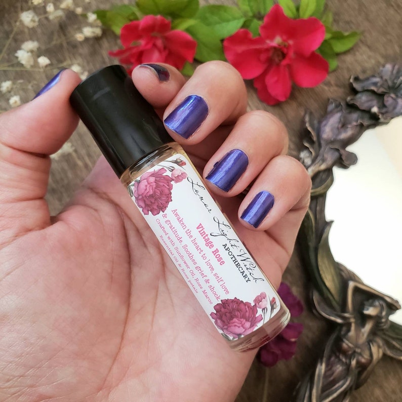Op de afbeelding: Een glazen flesje vintage rozen etherische olie met een bloemmotief en de tekst "Lyst Witch Apothecary Vintage Rose Awaken the heart to love, self love & gratitude. Soothes grief & shock. Crafted with Sunflower Oil, Rose Marigold"
