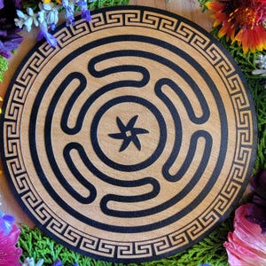 May include: A round wooden labyrinth with a black spiral design and a star in the center. The outer edge features a black Greek key pattern. The wood has a light brown color. The labyrinth is surrounded by colorful flowers.