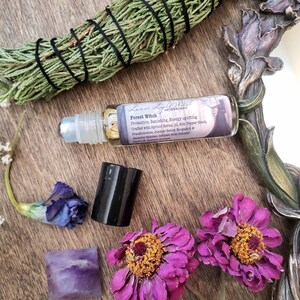 Pu&ograve; includere: Un flacone di vetro con rullo con un'etichetta nera che recita "Forest Witch Apothecary" e elenca gli ingredienti: olio di nocciolo di albicocca, pepe nero, incenso, bacca di ginepro, bergamotto, gelsomino sambac. Il flacone &egrave; appoggiato su una superficie di legno con fiori viola e un pezzo di ametista.