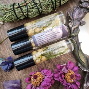 Pu&ograve; includere: Tre flaconi di vetro con rullo pieni di un liquido trasparente e prodotti botanici. I flaconi hanno tappi neri ed etichette che riportano "Forest Witch" e elencano gli ingredienti, tra cui olio di nocciolo di albicocca, pepe nero, incenso, bacca di ginepro, bergamotto e gelsomino sambac. I flaconi sono su una superficie di legno con fiori secchi e un pezzo di ametista.