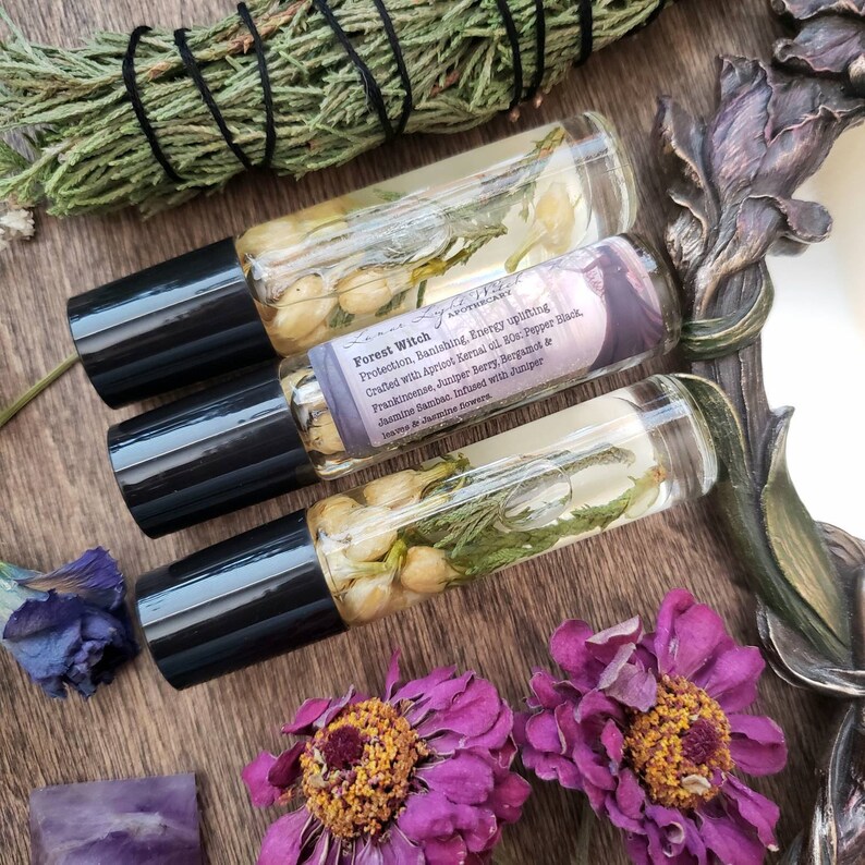 Pu&ograve; includere: Tre flaconi di vetro con rullo pieni di un liquido trasparente e prodotti botanici. I flaconi hanno tappi neri ed etichette che riportano "Forest Witch" e elencano gli ingredienti, tra cui olio di nocciolo di albicocca, pepe nero, incenso, bacca di ginepro, bergamotto e gelsomino sambac. I flaconi sono su una superficie di legno con fiori secchi e un pezzo di ametista.