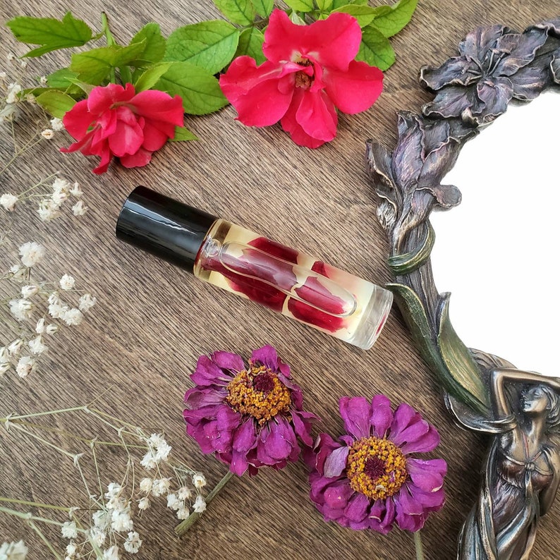 Op de afbeelding: Een transparante glazen rollerfles met een zwarte dop, gevuld met een gouden vloeistof en rode rozenblaadjes. De fles ligt op een houten oppervlak met gedroogde bloemen en een decoratieve spiegel.