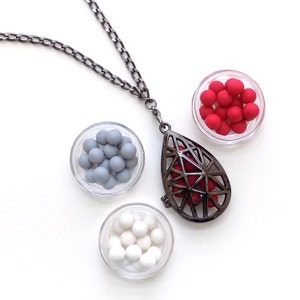 May include: A black chain necklace with a teardrop-shaped pendant. The pendant is made of metal and has a geometric design. There are three small bowls filled with red, white, and gray beads.