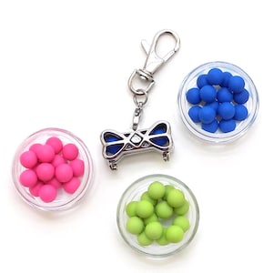 Puede incluir: Un charm plateado con un diseño de lazo azul y plateado, unido a un llavero plateado. Tres pequeños platos de vidrio contienen cuentas de colores: rosa, azul y verde.