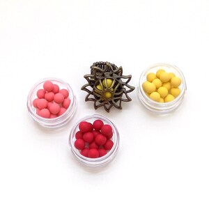 May include: Three small clear containers with lids, each filled with colorful round beads. The containers are arranged in a triangle shape. A decorative metal sun-shaped ring is in the center of the triangle.