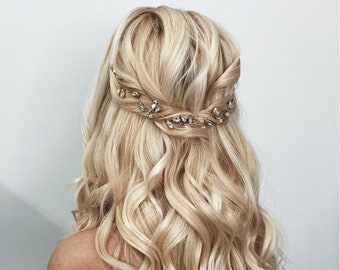 May include: A woman with long, blonde, wavy hair wearing a white off-the-shoulder top. Her hair is styled with a delicate gold and crystal hairpiece.