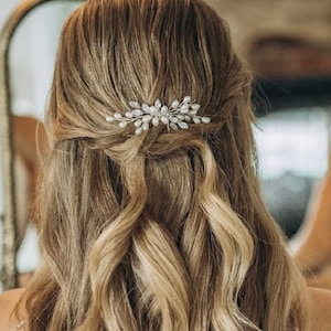 Könnte beinhalten: Lange, wellige blonde Haare, die zu einer halben Hochsteckfrisur gestylt sind, verziert mit einer silbernen Haarspange. Das Haar fällt kaskadenartig über den Rücken. Die Frau trägt ein weißes Kleid. Ein Spiegel ist im Hintergrund.