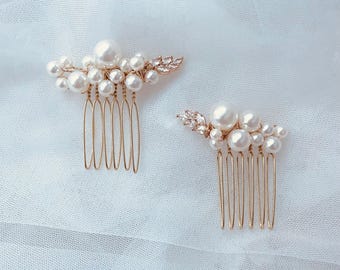 Puede incluir: Dos peinetas doradas, cada una adornada con perlas de imitación y detalles de cristal. Las peinetas presentan un grupo de perlas y un diseño de hojas decorativas. Las peinetas están sobre un fondo blanco texturizado.