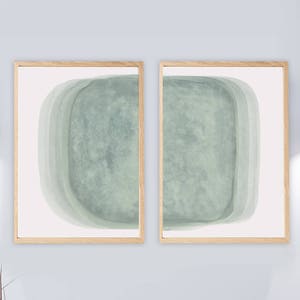 May include: Two framed abstract art prints with a light green watercolor design. The prints are hung on a light gray wall above a white dresser with copper drawer pulls.