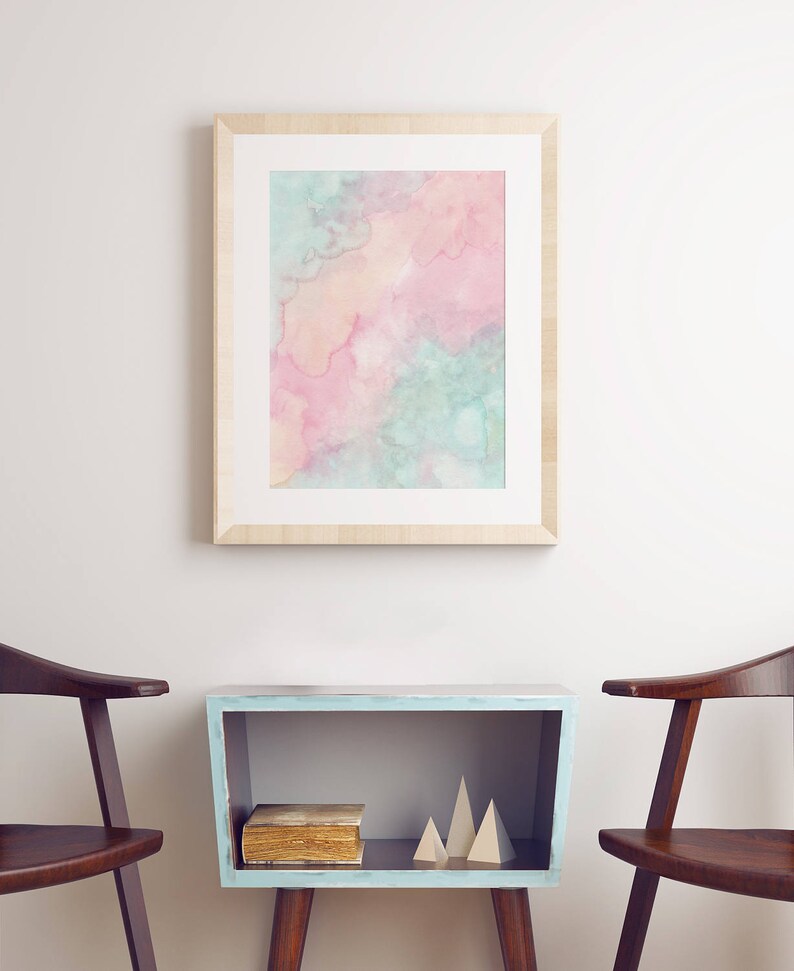 Puede incluir: Una pintura de acuarela enmarcada con tonos rosados, azules y amarillos cuelga sobre una mesa auxiliar azul claro con un libro y formas geom&eacute;tricas de madera. Dos sillas de madera flanquean la mesa.