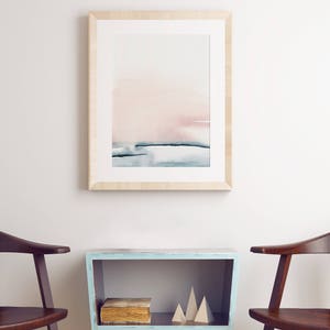 May include: A framed watercolor painting of a pink and blue abstract landscape hangs above a light blue shelf with a book and wooden geometric shapes. Two wooden chairs are on either side of the shelf.