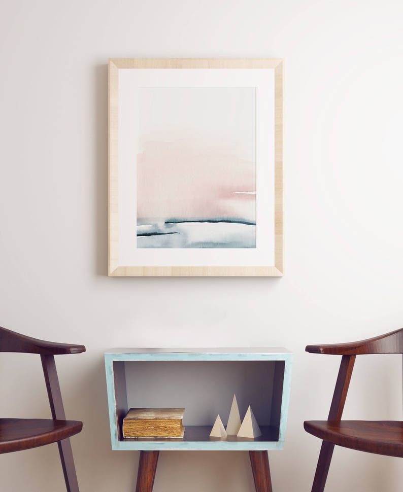 May include: A framed watercolor painting of a pink and blue abstract landscape hangs above a light blue shelf with a book and wooden geometric shapes. Two wooden chairs are on either side of the shelf.