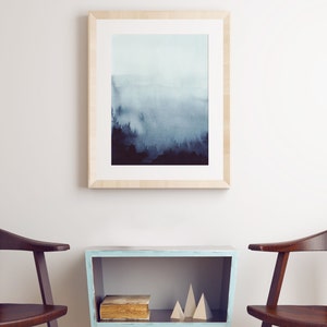 May include: A framed watercolor painting of a foggy forest scene in shades of blue and gray, hanging on a white wall above a light blue side table with two wooden chairs.