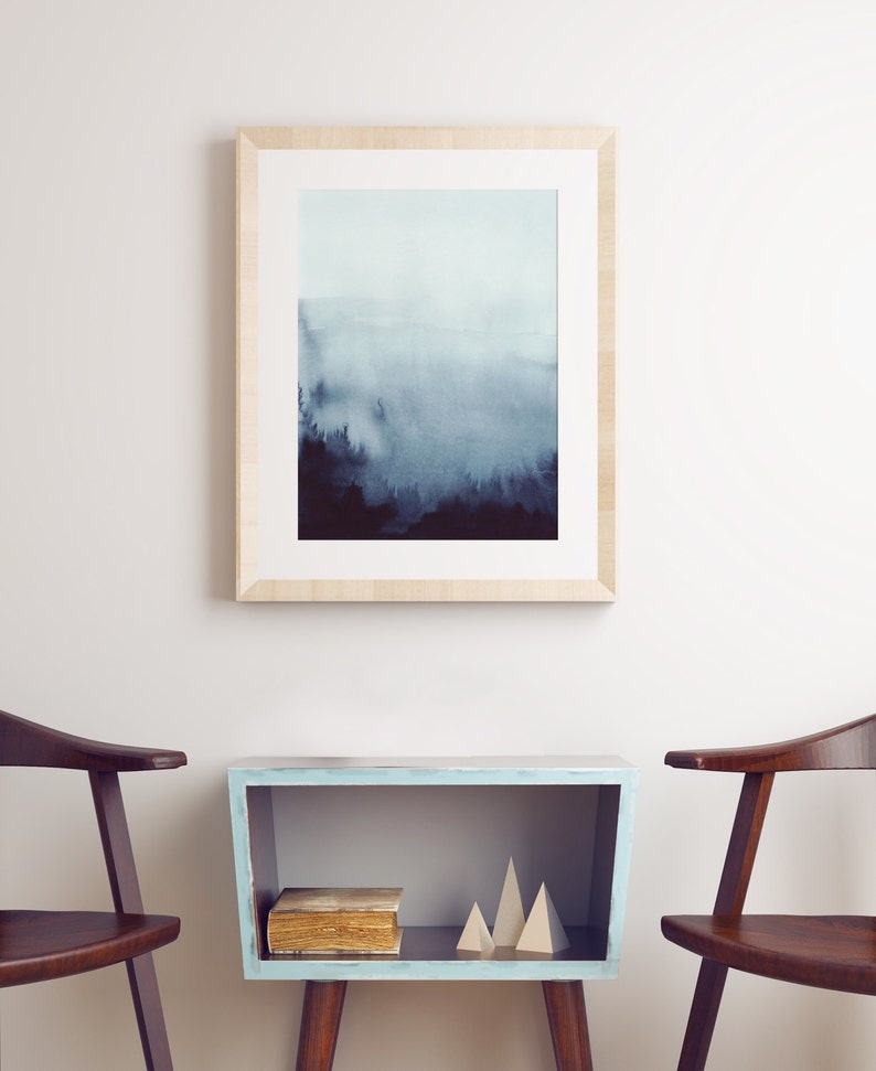 May include: A framed watercolor painting of a foggy forest scene in shades of blue and gray, hanging on a white wall above a light blue side table with two wooden chairs.