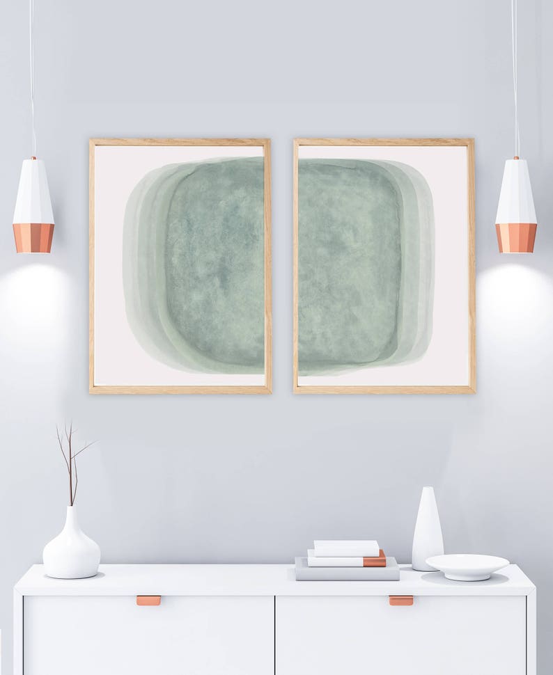 May include: Two framed abstract art prints with a light green watercolor design. The prints are hung on a light gray wall above a white dresser with copper drawer pulls.