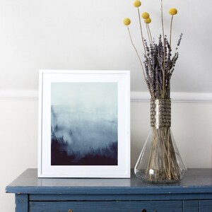 May include: A white framed print of a watercolor painting depicting a foggy forest scene in shades of blue and purple. The print is displayed on a blue dresser with a vase of dried lavender and yellow billy balls.