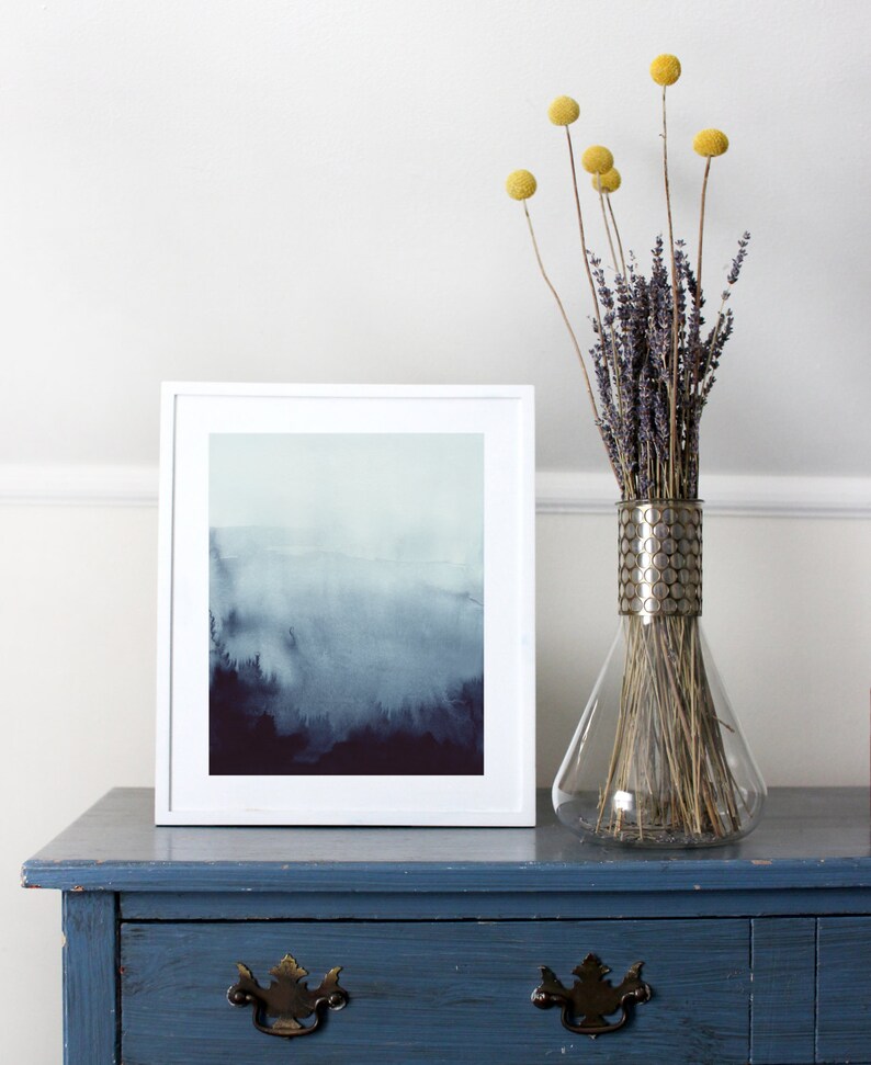 May include: A white framed print of a watercolor painting depicting a foggy forest scene in shades of blue and purple. The print is displayed on a blue dresser with a vase of dried lavender and yellow billy balls.