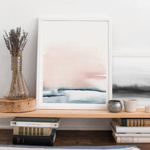 May include: A framed watercolor painting with a pink and blue abstract design. The painting is hanging on a shelf above a stack of books.