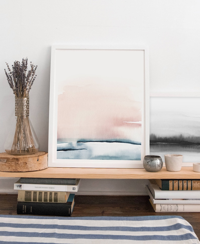 May include: A framed watercolor painting with a pink and blue abstract design. The painting is hanging on a shelf above a stack of books.