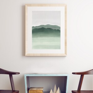 May include: A framed watercolor painting of a green mountain range with a light blue sky. The painting is hanging on a white wall above a light blue shelf with a book and geometric shapes.