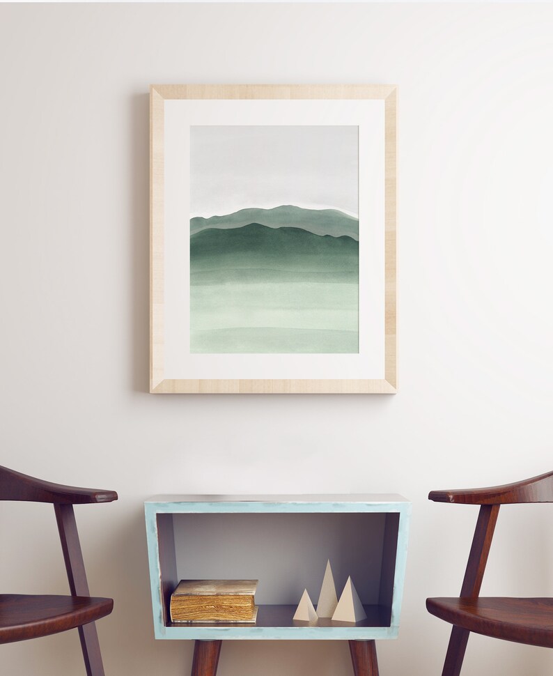 May include: A framed watercolor painting of a green mountain range with a light blue sky. The painting is hanging on a white wall above a light blue shelf with a book and geometric shapes.