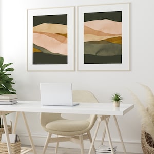 May include: Two framed abstract landscape prints with a muted color palette of green, brown, and pink. The prints are hanging on a white wall above a white desk with a laptop, a chair, and a potted plant.