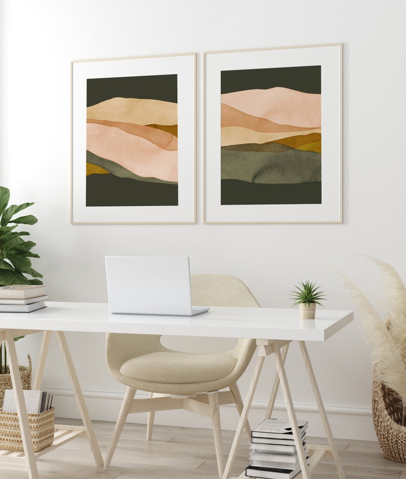 May include: Two framed abstract landscape prints with a muted color palette of green, brown, and pink. The prints are hanging on a white wall above a white desk with a laptop, a chair, and a potted plant.