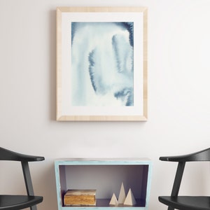 May include: A light blue wooden shelf with a book and two wooden geometric shapes on top. The shelf is sitting between two black chairs against a white wall. A framed watercolour painting of blue and white abstract shapes hangs on the wall above the shelf.