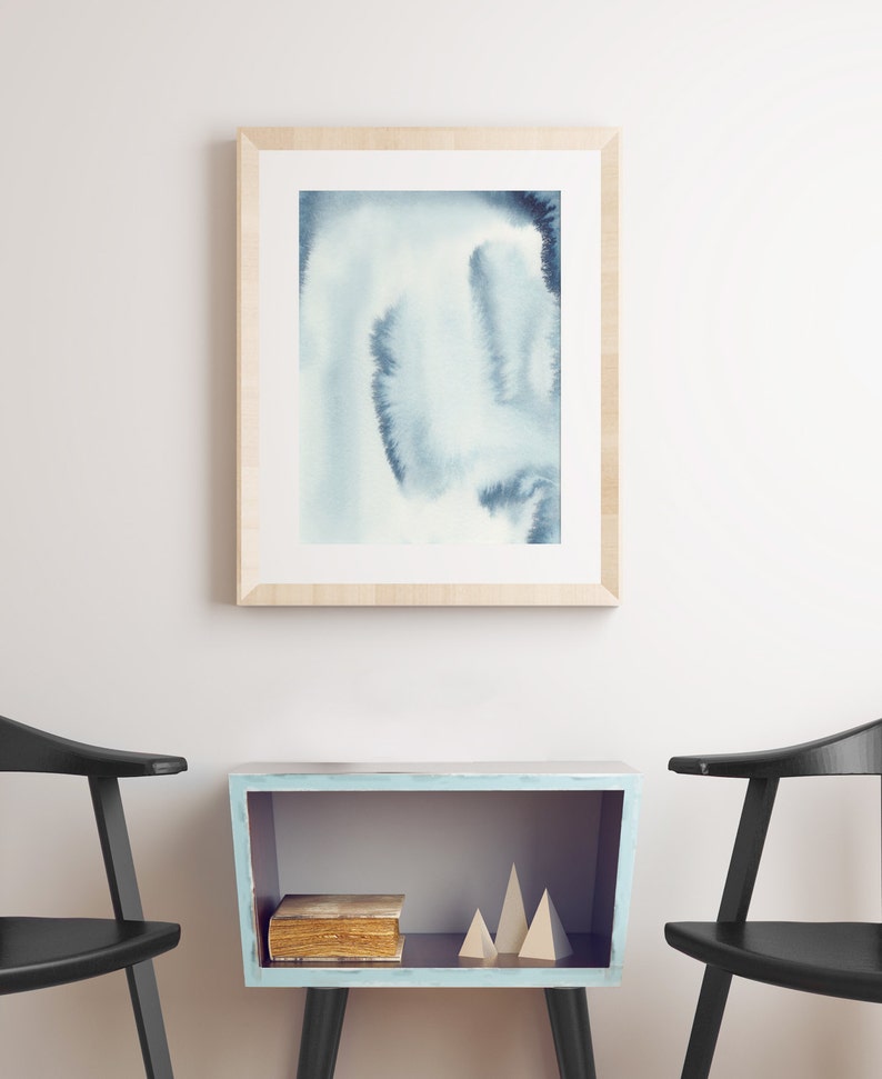 May include: A light blue wooden shelf with a book and two wooden geometric shapes on top. The shelf is sitting between two black chairs against a white wall. A framed watercolour painting of blue and white abstract shapes hangs on the wall above the shelf.