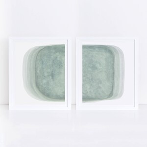 May include: Two framed abstract paintings with a light green, watercolor-like design. The paintings are in white frames and are hung on a white wall.