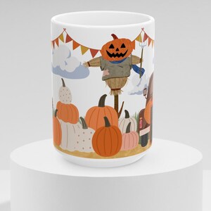 Puede incluir: Taza de cerámica blanca con un diseño de escena de cosecha otoñal. El diseño incluye un espantapájaros con forma de calabaza sonriente, calabazas y banderines festivos. La taza cilíndrica está sobre un pedestal blanco.