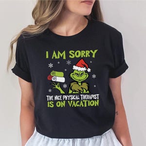 May include: Black t-shirt with a green Grinch wearing a Santa hat and holding a stack of green towels. The text on the shirt reads "I am sorry the nice physical therapist is on vacation."