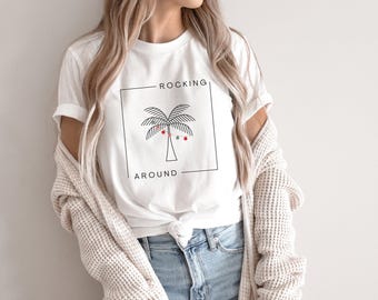 May include: White t-shirt with a black square containing a palm tree illustration and the words "ROCKING AROUND". The palm tree has red and green accents. The shirt is paired with blue jeans and a cream-colored cardigan.