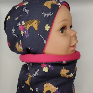 Könnte beinhalten: Ein dunkelblaues Mützen- und Halstuch-Set mit einem rosa Fleecefutter. Der Stoff hat ein sich wiederholendes Muster aus braunen Pferden, Blumen und Blättern.