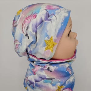 Könnte beinhalten: Ein Kinder-Set aus Mütze und Halstuch mit Einhorn-Motiv. Der Stoff ist mit Einhörnern, Wolken und goldenen Glitzersternen bedruckt. Das Set ist überwiegend blau, rosa und weiß.