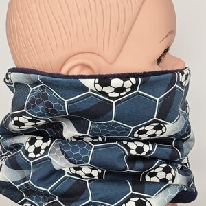 Könnte beinhalten: Ein blau-weißes Halstuch mit Fußballmuster. Das Halstuch hat ein sich wiederholendes Muster aus Fußbällen in einem sechseckigen Design. Die Innenseite ist aus dunkelblauem Plüschmaterial. Das Halstuch soll den Hals warm halten.