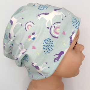 Könnte beinhalten: Ein hellblauer Beanie-Hut mit einem sich wiederholenden Muster aus weißen Einhörnern, Regenbogen und farbenfrohen Akzenten. Der Hut ist aus weichem Stoff gefertigt und ist so konzipiert, dass er bequem sitzt. Dieser Hut ist perfekt, um jedem Outfit einen Hauch von Laune zu verleihen.