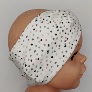 Könnte beinhalten: Ein weißes Stirnband mit einem grauen, schwarzen und rosafarbenen Tupfenmuster. Das Stirnband ist aus weichem, dehnbarem Stoff gefertigt und soll als Turban getragen werden.