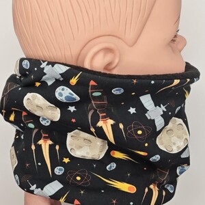 Könnte beinhalten: Schwarzer Halswärmer mit Weltraum-Motiv. Das Design umfasst Raketen, Planeten, Sterne und Satelliten. Der Halswärmer ist aus einem weichen Material gefertigt und soll den Träger warm halten. Der Halswärmer wird an einer Puppe gezeigt.