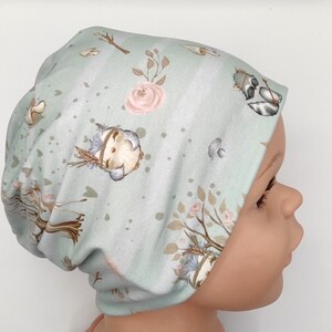 Könnte beinhalten: Eine hellblaue Baby-Mütze mit einem Waldtier-Druck. Die Mütze zeigt einen kleinen Vogel, einen Waschbären, Pilze und Blumen.