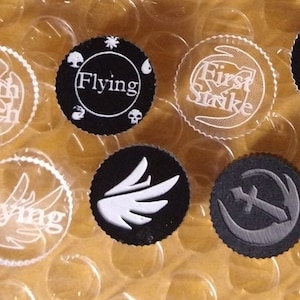 May include: A collection of circular game tokens with various symbols and text. The tokens feature phrases like "Death Touch," "Flying," and "First Strike." The designs include wings, swords, and other stylized graphics, in white and gray on black backgrounds.