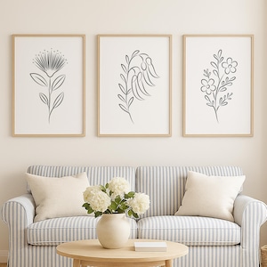 Op de afbeelding: Drie ingelijste botanische kunstprints met zwarte lijntekeningen van bloemen en bladeren. Ze hangen boven een blauw-witte gestreepte bank met crèmekleurige kussens. Een ronde houten salontafel en een rieten mand maken de scène compleet.