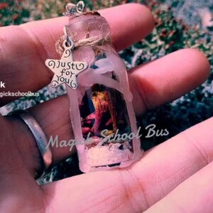 Puede incluir: Un collar con una pequeña botella de vidrio y un dije de estrella plateada que dice "Just for you". La botella está llena de flores secas y sellada con cera. El collar está en una mano.