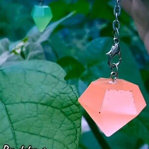 Może przedstawiać: Różowy, diamentowy wisiorek z żywicy wiszący na srebrnym łańcuszku z karabińczykiem. Wisiorek jest przezroczysty i lekko błyszczy.